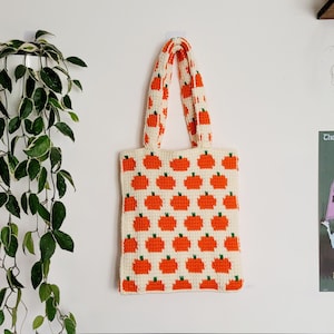 Könnte beinhalten: Eine weiße gehäkelte Tragetasche mit orangefarbenen Kürbissen. Die Tasche hat zwei Henkel und hängt an einer weißen Wand.
