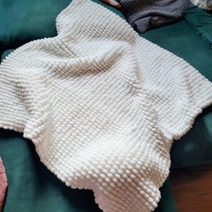 Könnte beinhalten: Eine weiße Decke mit einem strukturierten, blasenartigen Muster. Die Decke ist gefaltet und liegt auf einer grünen Oberfläche, wodurch ihr weiches, plüschiges Aussehen hervorgehoben wird. Das Design der Decke deutet auf ein gemütliches Heimtextil hin.