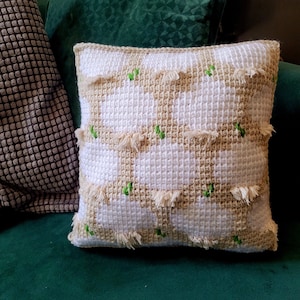 May include: A white and beige crocheted pillow with green accents. The pillow has a geometric pattern and fringe details.