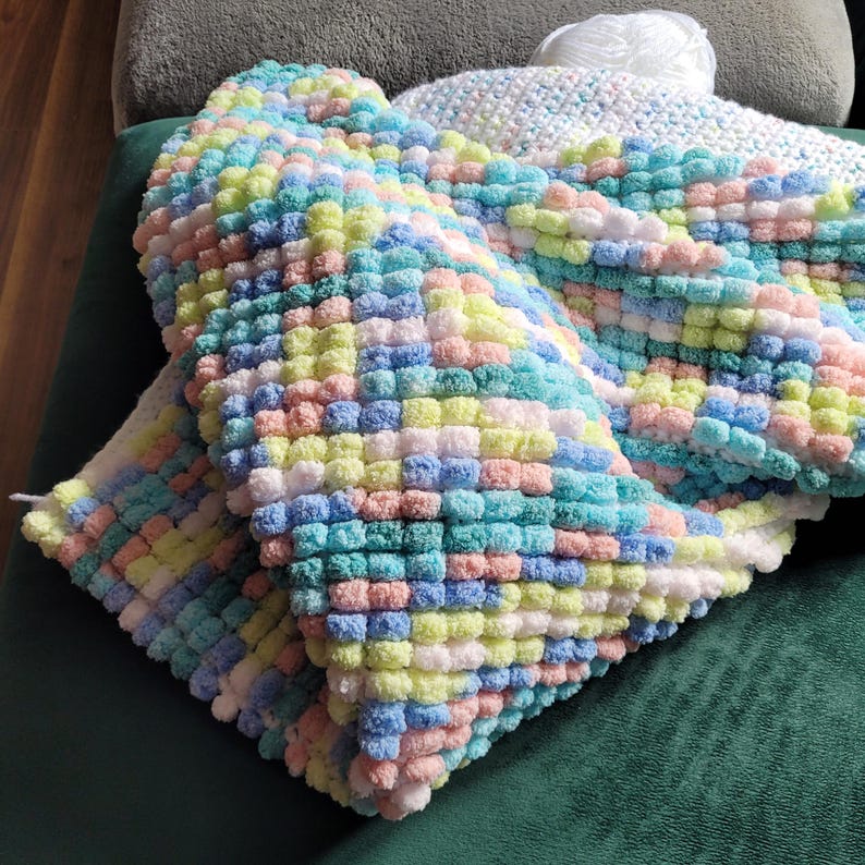 May include: A colorful knitted blanket with a textured, puffy design. The blanket is made with a variety of pastel colors, including pink, blue, yellow, and white.