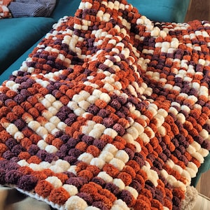 Könnte beinhalten: Eine weiche, plüschige Decke aus Pompons in den Farben Weiß, Braun und Orange. Die Decke ist diagonal angeordnet und erzeugt ein strukturiertes und optisch ansprechendes Design.