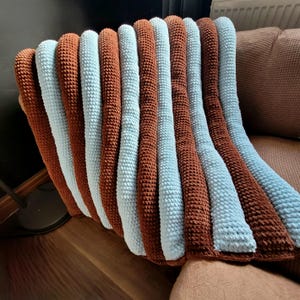 Pode incluir: Uma manta feita à mão em crochê com listras verticais alternadas de fio castanho macio e azul claro. O tecido tem uma aparência espessa, felpuda e texturizada criada pelo padrão de pontos repetidos, formando cristas arredondadas.
