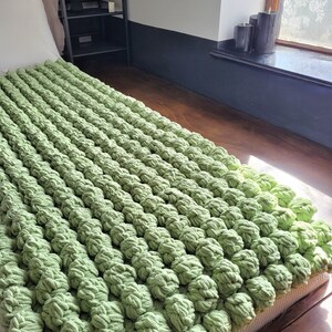 Op de afbeelding: Een groene gehaakte deken met een textuur, een bobbelig oppervlak. De deken is gemaakt van dik garen en voelt zacht en comfortabel aan.