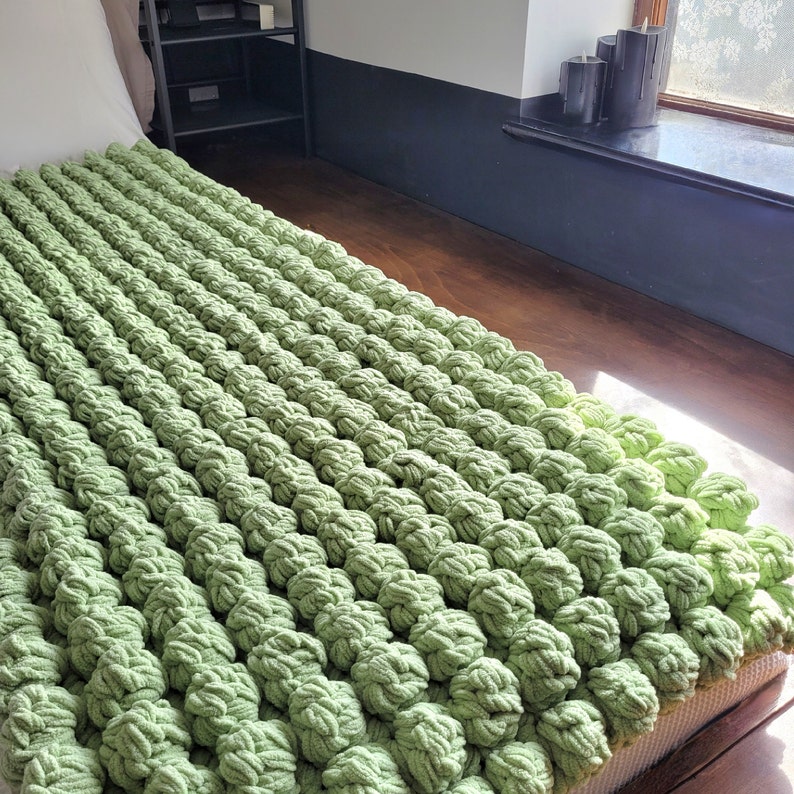 Op de afbeelding: Een groene gehaakte deken met een textuur, een bobbelig oppervlak. De deken is gemaakt van dik garen en voelt zacht en comfortabel aan.