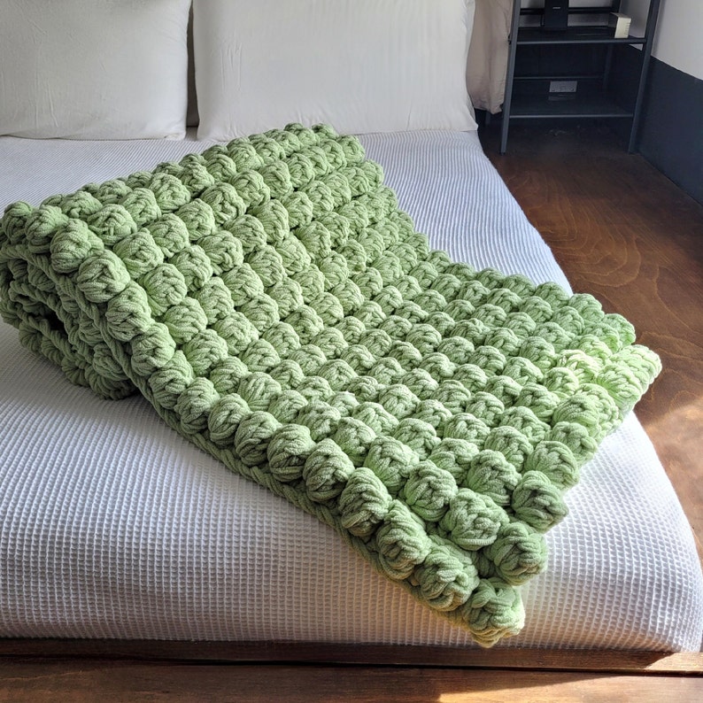 Op de afbeelding: Een zacht, groen gebreid deken met een textuur, bubbelachtig ontwerp. Het deken is gedrapeerd over een wit beddengoed.