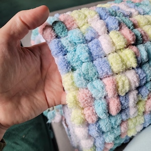 May include: A soft, colorful blanket made with fluffy yarn. The blanket is made in a variety of pastel colors, including pink, blue, green, and white. The blanket is made with a textured stitch that creates a pom-pom effect.