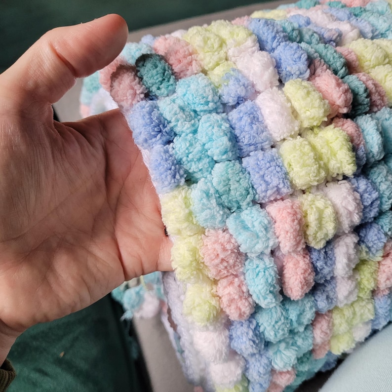 May include: A soft, colorful blanket made with fluffy yarn. The blanket is made in a variety of pastel colors, including pink, blue, green, and white. The blanket is made with a textured stitch that creates a pom-pom effect.