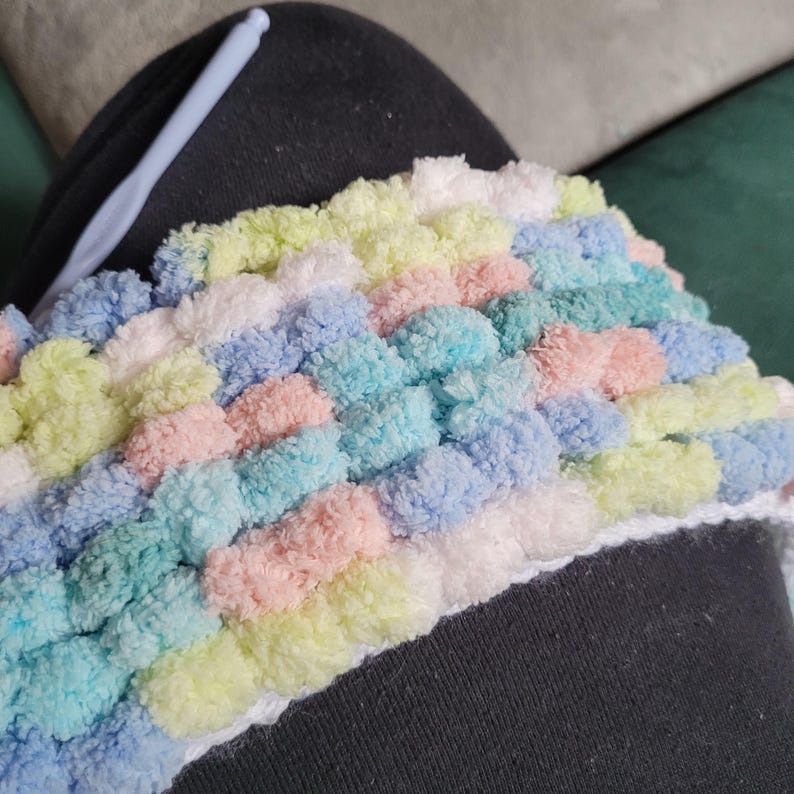 May include: A close-up of a colorful crocheted blanket with a variety of pastel colors, including pink, blue, yellow, and white. The blanket is made with a fluffy yarn and has a textured surface.
