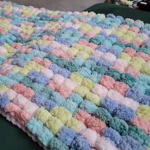 May include: A colorful knitted blanket with a pom-pom texture in shades of pink, blue, green, and white.