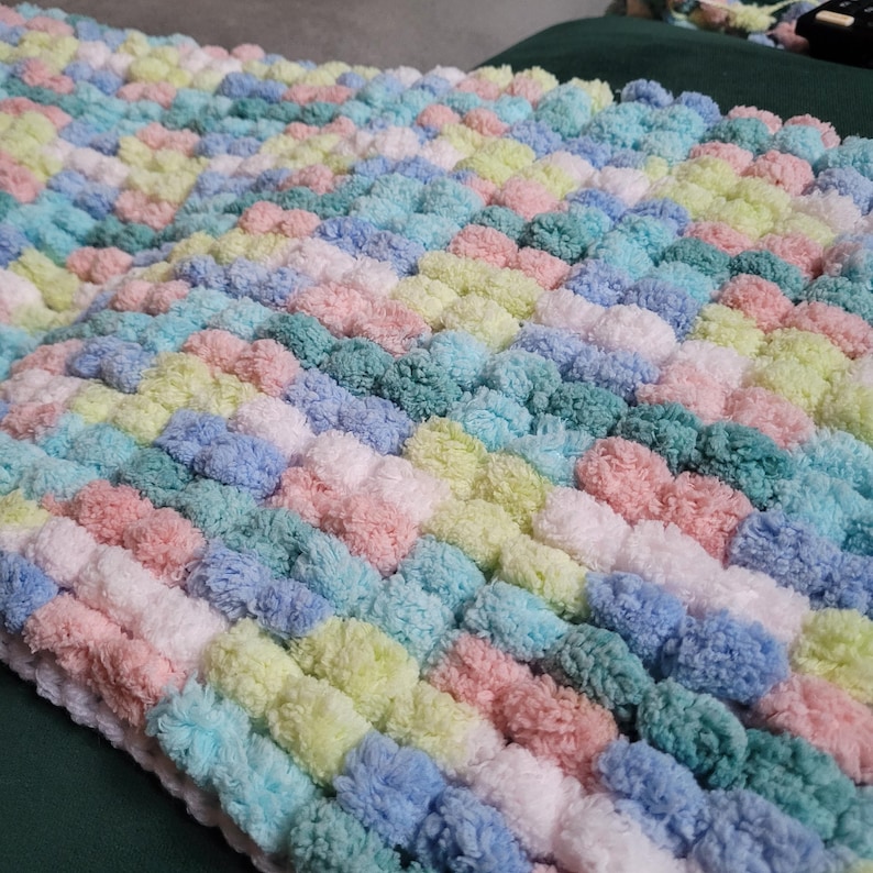 May include: A colorful knitted blanket with a pom-pom texture in shades of pink, blue, green, and white.