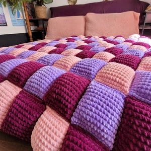 Könnte beinhalten: Eine flauschige, grob gestrickte Decke mit einem sich wiederholenden Gittermuster aus weichen, erhabenen Quadraten in abwechselnden Farbtönen von Hellrosa, Lavendel und tiefem Magenta. Das strukturierte Garn sorgt für eine dicke, gepolsterte Oberfläche.