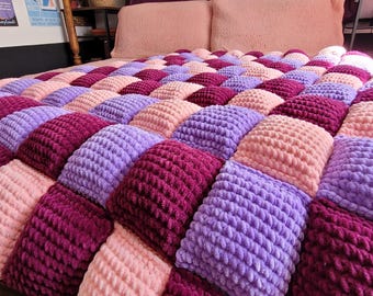 Puede incluir: Una manta de punto grueso y afelpado con un patrón de cuadrícula repetitivo de cuadrados suaves y elevados en tonos alternos de rosa claro, lavanda y magenta intenso. El hilo texturizado crea una superficie gruesa y acolchada uniforme.