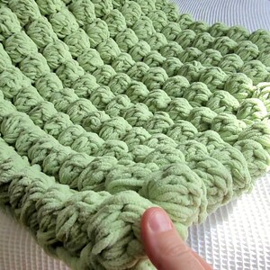 Op de afbeelding: Een zacht, groen gebreid deken met een textuur, een bolletjes ontwerp. Het deken is gemaakt van dik garen en heeft een pluche, zachte uitstraling.