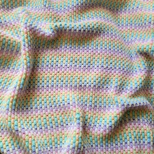 Könnte beinhalten: Nahaufnahme einer pastellfarbenen Häkeldecke. Die Decke weist ein sich wiederholendes Muster aus vertikalen und horizontalen Stichen in Rosa-, Gelb-, Blau- und Grüntönen auf. Der weiche, strukturierte Stoff ist drapiert und erzeugt Falten und Schatten.