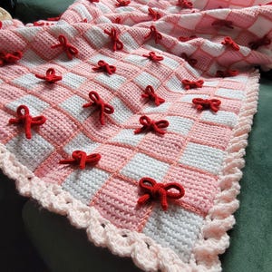May include: A handmade crochet blanket with a pink and white checkered pattern. The blanket is adorned with numerous red bows and has a scalloped edge. The blanket is made of soft yarn and is perfect for a baby or child.