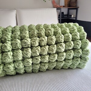 Op de afbeelding: Een zachte, groene gebreide deken met een unieke bubbelstructuur. De deken is gemaakt van garen en heeft een pluche, knusse uitstraling.