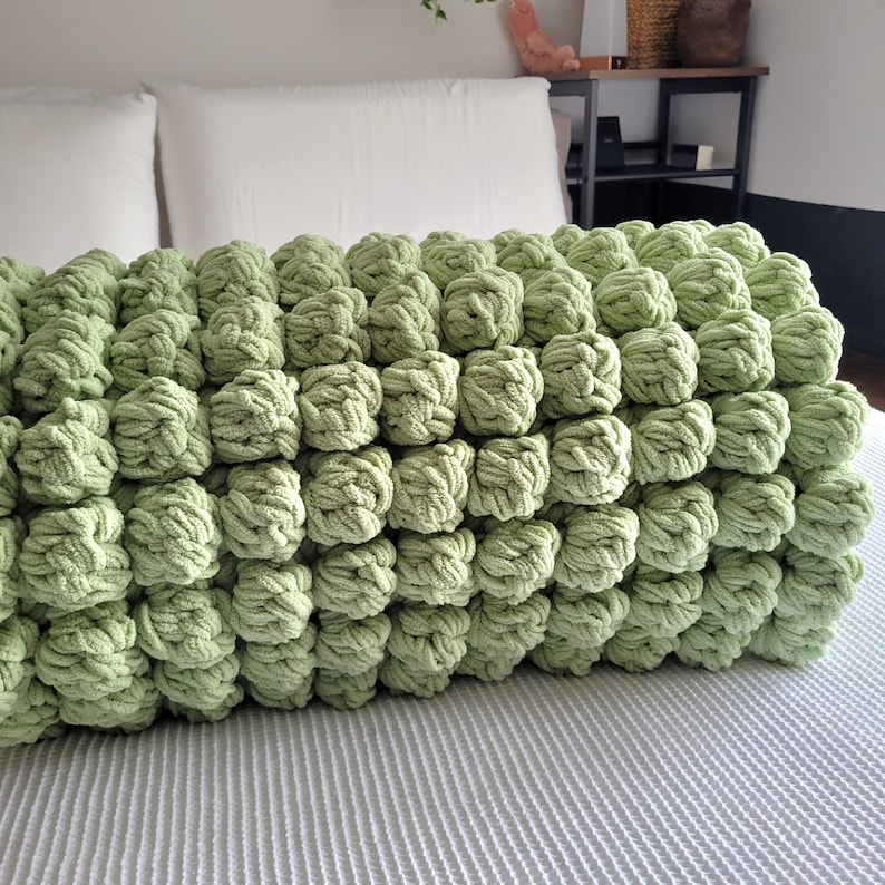 Op de afbeelding: Een zachte, groene gebreide deken met een unieke bubbelstructuur. De deken is gemaakt van garen en heeft een pluche, knusse uitstraling.