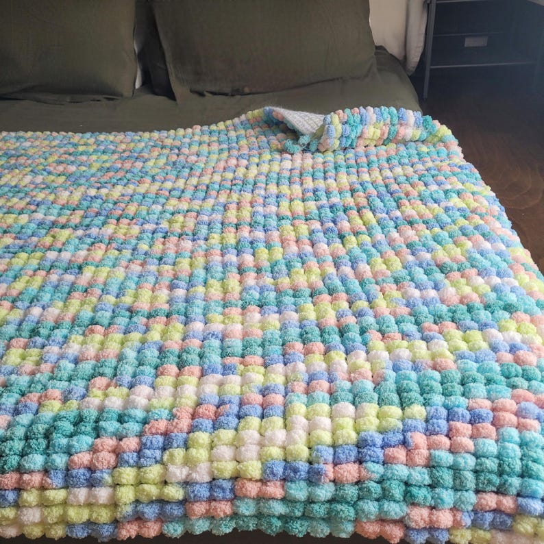 May include: A soft, colorful knitted blanket with a pom-pom texture. The blanket is made in a variety of pastel colors, including pink, blue, green, and white.