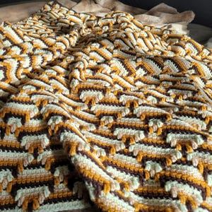 May include: A handmade crochet blanket with a geometric pattern. The blanket features a repeating design in shades of brown, cream, yellow, and pale green. The textured surface is soft and inviting, perfect for adding warmth and style to any space.