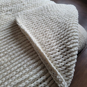 Könnte beinhalten: Eine weiße Häkeldecke mit einem dicken, strukturierten Design. Die Decke ist in der Hälfte gefaltet und zeigt die weiche, flauschige Textur.