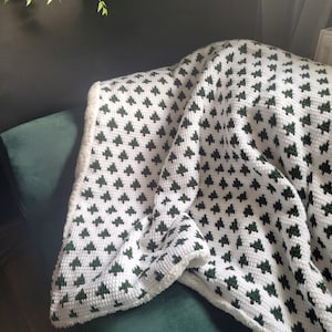 Könnte beinhalten: Eine weiße Häkeldecke mit einem grünen Baummuster. Die Decke hat einen flauschigen weißen Rand.