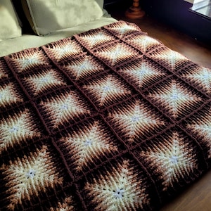 Könnte beinhalten: Eine handgefertigte Häkeldecke mit Sternmuster. Die Decke zeigt ein sich wiederholendes Muster aus cremefarbenen und braunen Sternformen vor einem dunkelbraunen Hintergrund. Die Decke wird auf einem Bett mit grünen Kissen präsentiert.