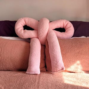 Pode incluir: Uma almofada grande, cor-de-rosa clara, tricotada à mão em forma de laço. A almofada apresenta uma construção de fio grosso e texturizado com um nó central e duas pontas longas e pendentes que caem sobre a superfície das almofadas.