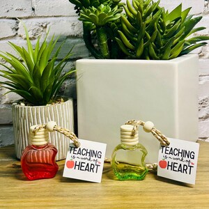 May include: Two small, apple-shaped bottles, one red and one green, with wooden tops and tags reading "TEACHING is a work of HEART." The bottles sit on a wooden surface, with plants in the background.