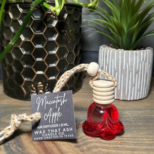 May include: A red glass air freshener with a wooden top and a woven cord. A black tag reads "Macintosh Apple" and "Air Diffuser 8 ML." The background includes potted plants.