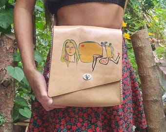 May include: A beige leather crossbody bag with a flap closure and a metal clasp. The bag features a hand-painted design of a woman's face and abstract shapes in yellow, red, and green.