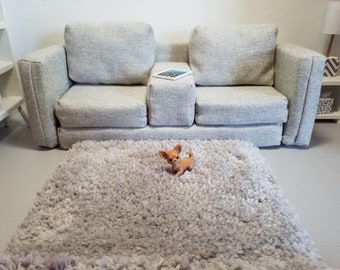 Könnte beinhalten: Ein hellgraues Sofa mit einer kleinen Hundefigur, die auf einem flauschigen grauen Teppich sitzt.
