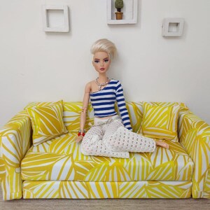 May include: A doll is seated on a yellow and white patterned sofa with matching throw pillows. The doll wears a blue and white striped top and white pants. The sofa is a miniature piece of furniture.