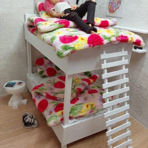 May include: A white wooden bunk bed set for dolls featuring a ladder, two mattresses, and two pillows covered in a fuzzy white fabric with a vibrant pink, red, and yellow floral pattern. A doll with pink hair lies on the top bunk wearing a white top and black jeans.