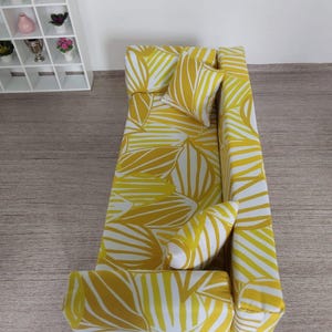 May include: A miniature yellow and white patterned sofa with two matching pillows. The sofa has a modern design with a palm leaf pattern. The sofa is placed on a wooden surface. A white shelf with small plants and decorative items is in the background.