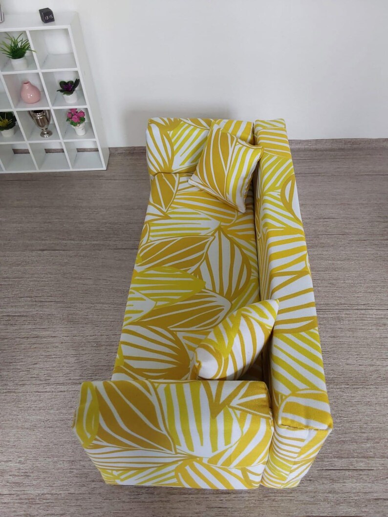 May include: A miniature yellow and white patterned sofa with two matching pillows. The sofa has a modern design with a palm leaf pattern. The sofa is placed on a wooden surface. A white shelf with small plants and decorative items is in the background.