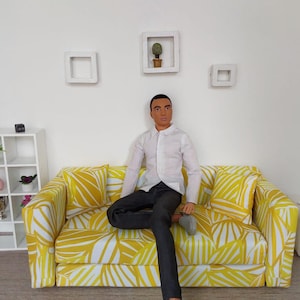May include: A doll dressed in a white button-down shirt and black pants sits on a yellow and white patterned sofa. The sofa is in a room with white walls, a white shelf, and decorative wall art.
