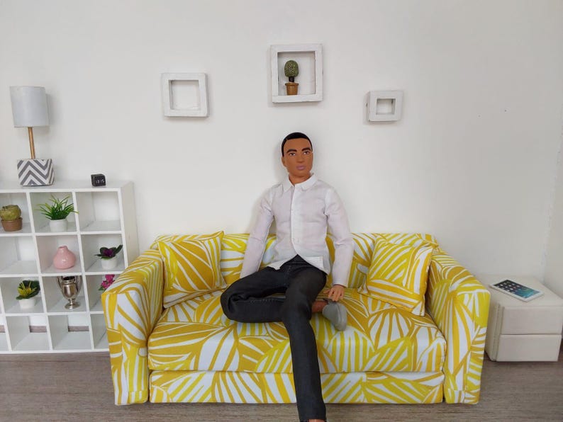 May include: A doll dressed in a white button-down shirt and black pants sits on a yellow and white patterned sofa. The sofa is in a room with white walls, a white shelf, and decorative wall art.