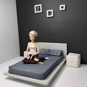 May include: A miniature bedroom set featuring a doll sitting on a gray bed with a dark blue mattress and pillows. The bed has a gray headboard and sits on a gray platform. Two white nightstands and three white square wall decorations complete the scene.