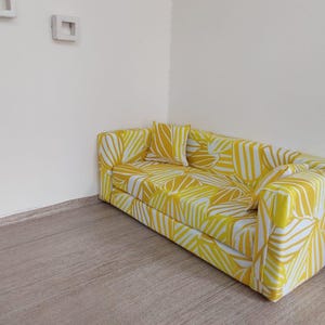 May include: A yellow and white patterned sofa with a matching pillow. The sofa is rectangular with a modern design. The pattern features a leaf-like design. The sofa is placed in a corner of a room with light-colored walls and a wooden floor.
