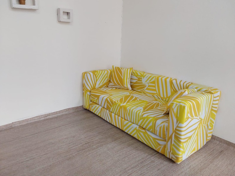 May include: A yellow and white patterned sofa with a matching pillow. The sofa is rectangular with a modern design. The pattern features a leaf-like design. The sofa is placed in a corner of a room with light-colored walls and a wooden floor.