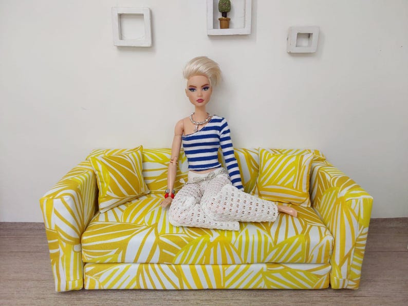 May include: A doll is seated on a yellow and white patterned sofa with matching throw pillows. The doll wears a blue and white striped top and white pants. The sofa is a miniature piece of furniture.