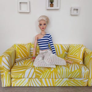 May include: A doll is seated on a miniature yellow sofa with a white leaf pattern. The doll wears a blue and white striped top, white pants, and a silver necklace. Two matching pillows are on the sofa.