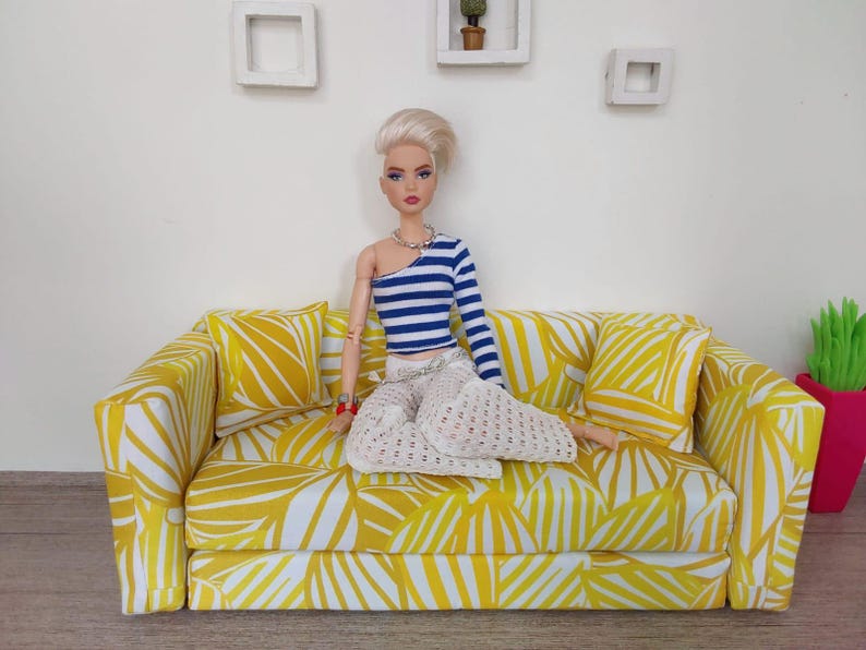 May include: A doll is seated on a miniature yellow sofa with a white leaf pattern. The doll wears a blue and white striped top, white pants, and a silver necklace. Two matching pillows are on the sofa.