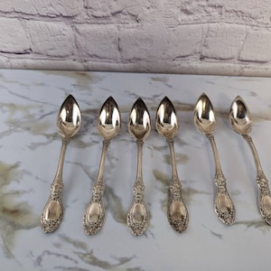 Puede incluir: Seis cucharas de plata con mangos adornados y una cabeza de cuchara redondeada. Las cucharas están dispuestas en fila sobre una superficie de mármol blanco.