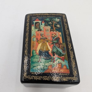Puede incluir: Una caja de madera negra con una escena pintada de dos personas con ropa tradicional rusa de pie frente a un castillo. La caja está decorada con bordes dorados y el texto "ПОДАРОК ИЗ РУССКОЙ СКАЗКИ" está escrito en la parte superior.