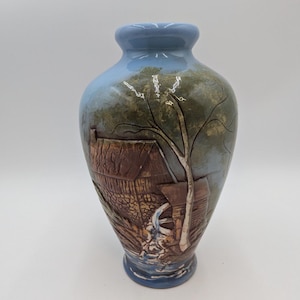 May include: A blue ceramic vase with a 3D scene of a rustic house and a water wheel. The scene is painted in brown, green, and white.