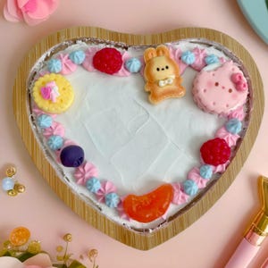 Könnte beinhalten: Ein herzförmiger Kuchen, der mit rosa, blauem und weißem Frosting verziert ist. Der Kuchen hat verschiedene Beläge, darunter ein Häschen, ein Hello-Kitty-Keks und verschiedene Süßigkeiten.