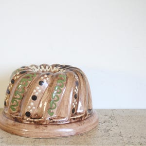 May include: A vintage brown ceramic bundt pan with a decorative design. The pan features a spiral shape with green and black accents. The base is round and the pan is suitable for baking cakes.