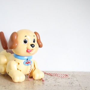 Pode incluir: Um cão de brinquedo de plástico com corpo cor de creme, orelhas castanhas e cauda castanha. O cão apresenta uma coleira azul brilhante com uma etiqueta vermelha e uma trela de corda às riscas vermelhas e brancas presa à frente.