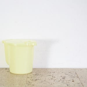 Può includere: Un'ampolla di plastica di colore giallo pallido con un coperchio. L'ampolla è appoggiata su una superficie di colore marrone chiaro.